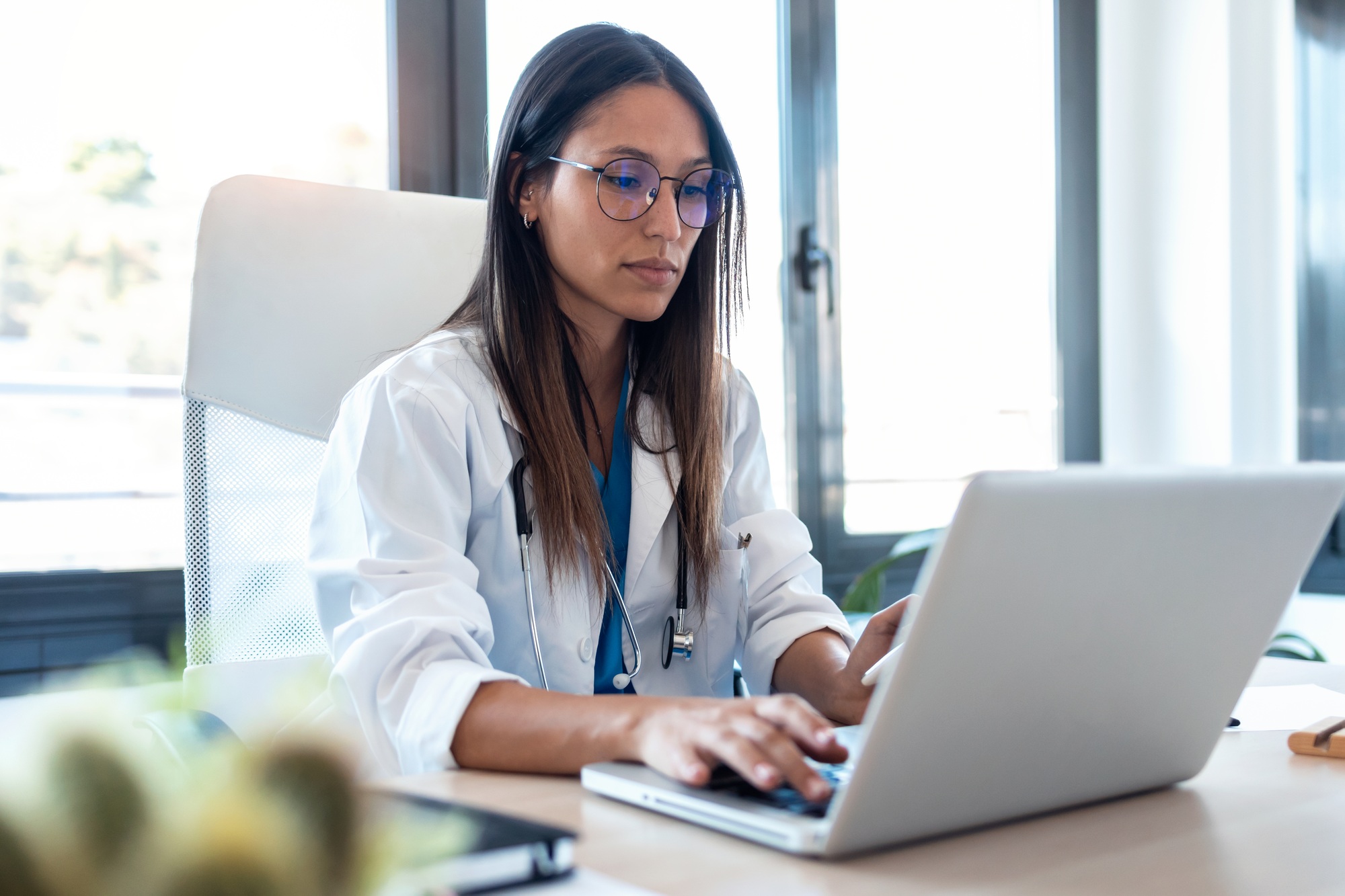 Confident young female doctor using her mobile phone while working on laptop in the consultation.